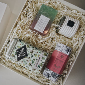 Close up of gift box contents which include Biodiversitea tea tin, magnolia pink hand cream, rectangle rose soap, and luxury travel candle. Beige crinkle paper fills the contents inside the box.