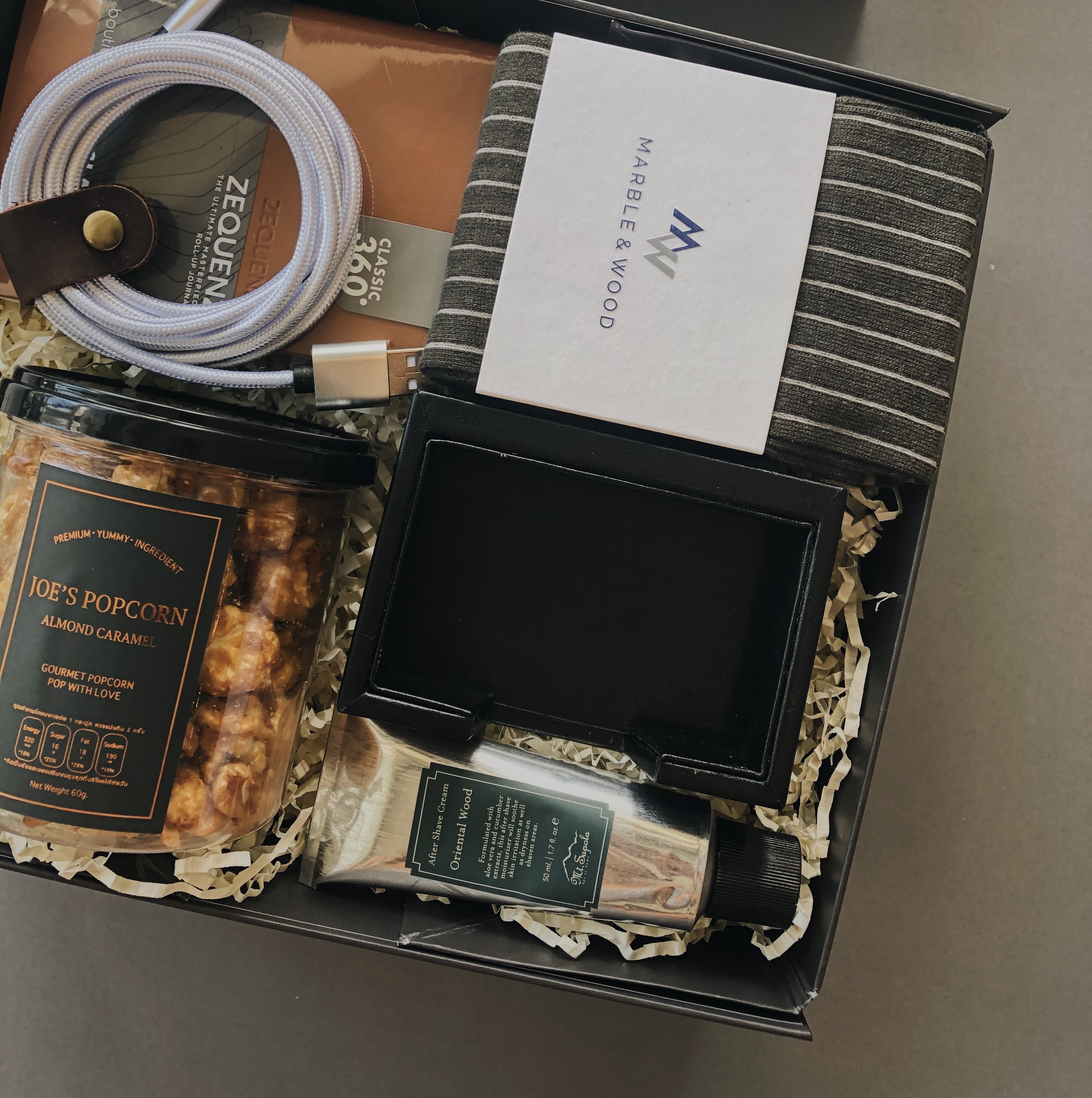 Close up of gift box filled with products inspired for a gentleman including luxury striped socks, brown A6 leather notebook, 2M phone charging cable, tub of almond caramel popcorn, black leather name card holder, silver tube of oriental wood aftershave.