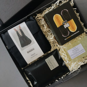 Dark grey rigid box with lid and black ribbon. Contents inside the box are stuffed with beige crinkle paper. Contents include luxury black box of shoe freshener shaped in a pill, black and white stripe reusable grocery bag, rectangle light yellow peppermint soap and black packet of spring tea hand sanitiser. 