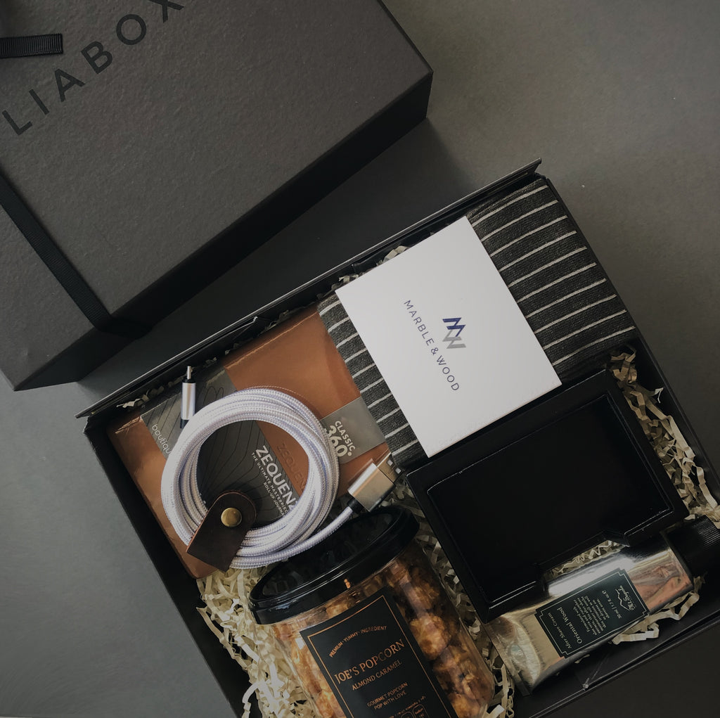 Dark grey rigid box with lid and black ribbon. Contents inside the box are stuffed with beige crinkle paper inspired for a gentleman and include luxury striped socks, brown A6 leather notebook, 2M phone charging cable, tub of almond caramel popcorn, black leather name card holder, silver tube of oriental wood aftershave.