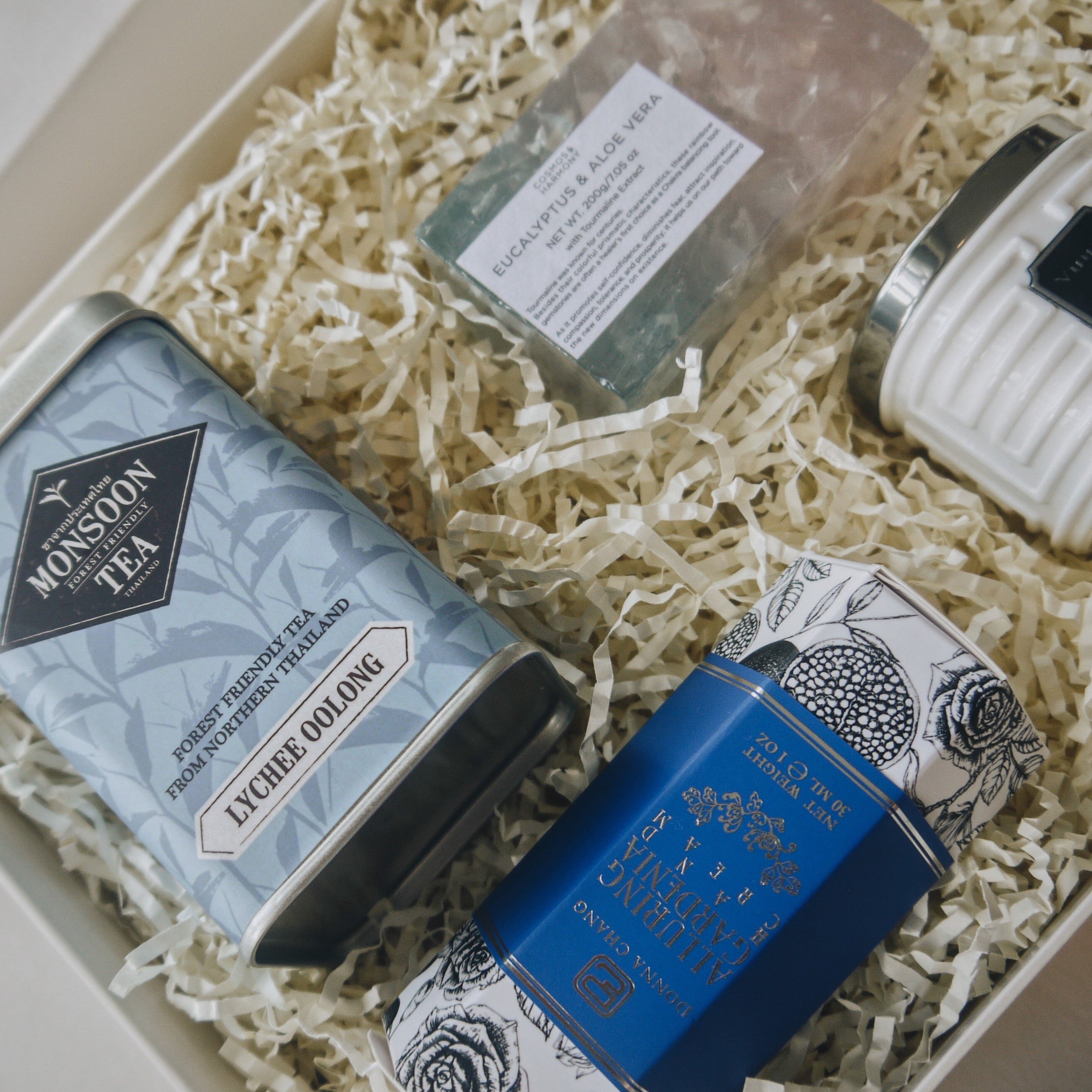 Close up of gift box contents which include lychee oolong tea tin, alluring gardenia hand cream, rectangle eucalyptus soap, and luxury travel candle. Beige crinkle paper fills the contents inside the box. 